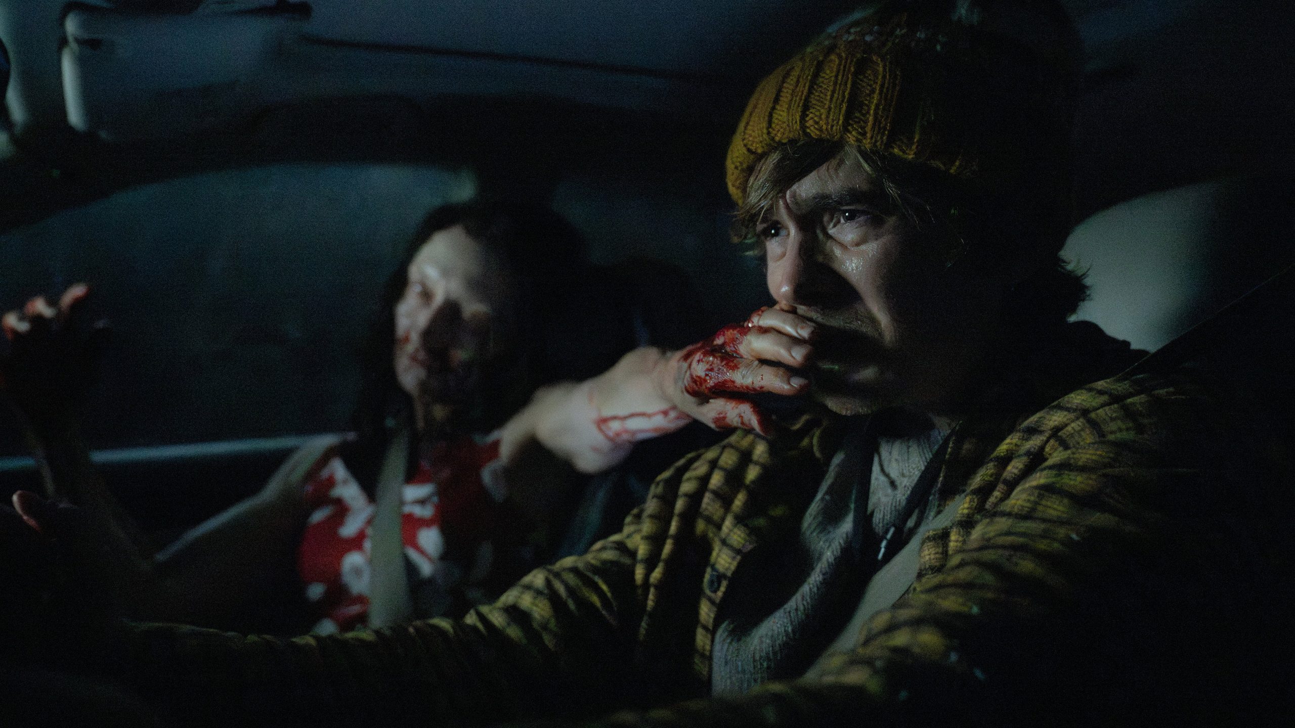 Bryan, a beleaguered medical courier in yellow winter gear, tries to drive despite the distraction of the zombie in the passenger seat of his car trying to grab his face.