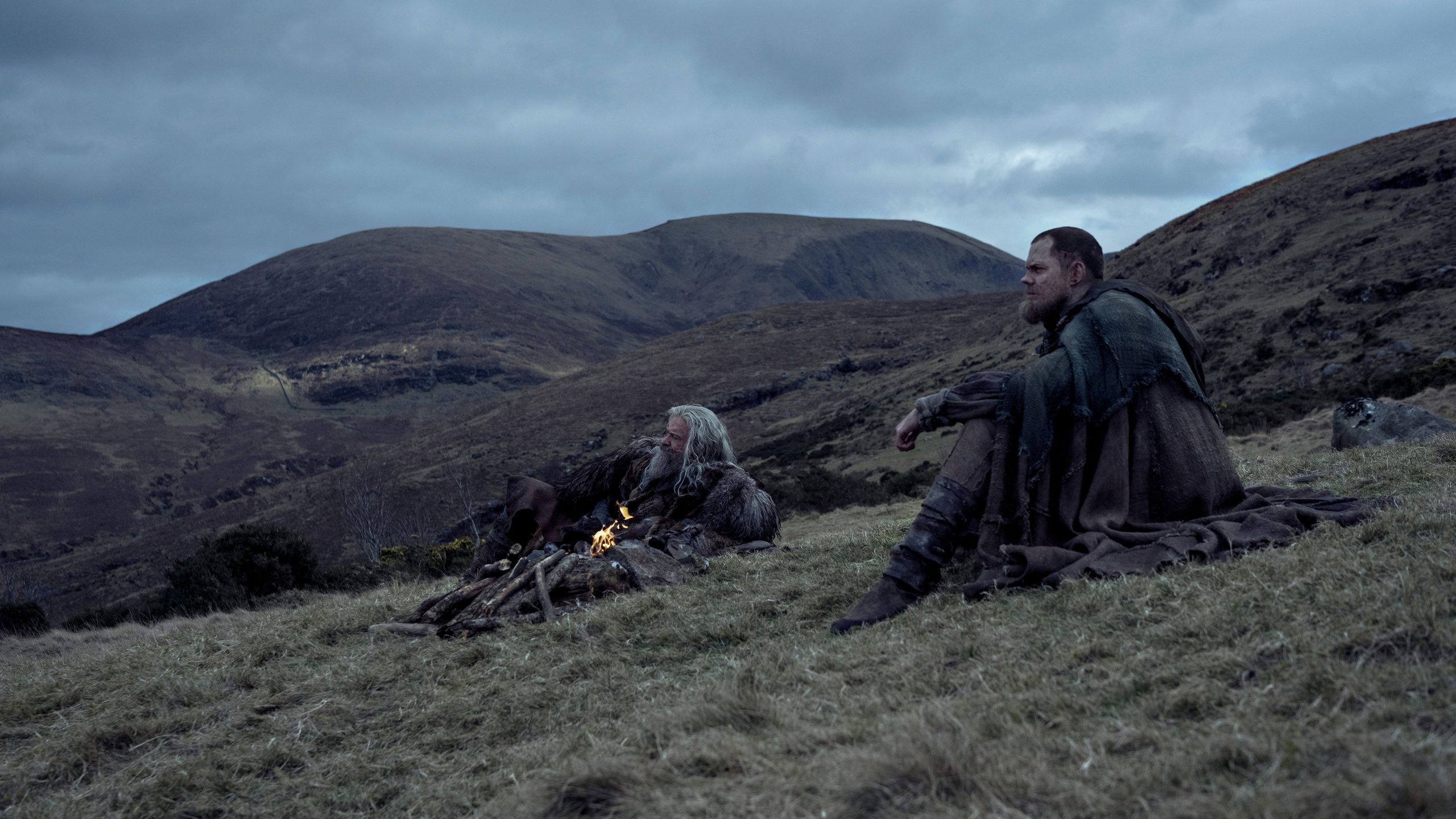 A wizened Robin Hood sits by the fire with Little John.